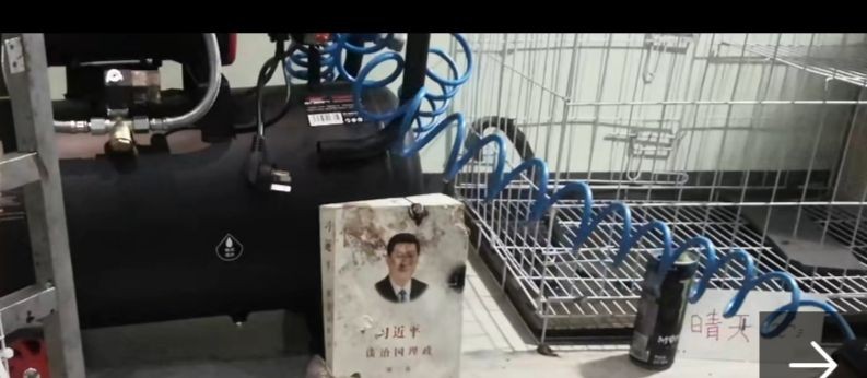 Equipment from Dark Lord's videos: a black air tank with a coiled blue hose in front of a wire cage, with a worn copy of Xi Jinping: The Governance of China propped against the cage and a small handwritten card reading 晴天 beside it.